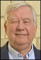 An older man with gray hair and a friendly smile, dressed in a light blue shirt and dark jacket against a neutral background.