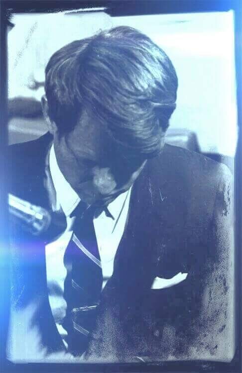 A man in a suit bows his head, focusing intently, with a microphone visible nearby, illuminated by a soft light.