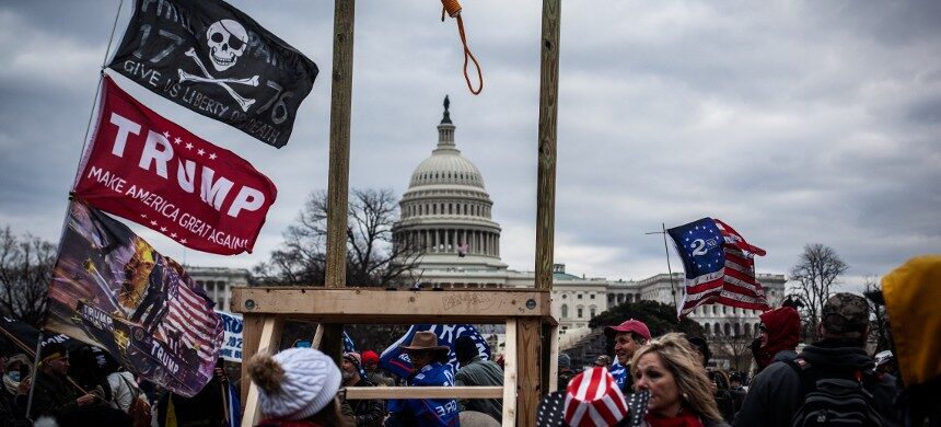 A crowd with flags gathers near a protest structure and the Capitol building, under a cloudy sky.