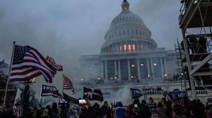 A large crowd gathers at the U.S. Capitol under smoke, with flags and banners visible amidst a tense atmosphere.