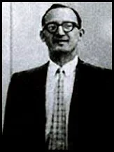 A man stands smiling, dressed in a black suit with a white shirt and plaid tie, wearing glasses in a monochrome photo.