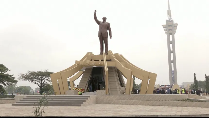 A towering statue stands atop a golden structure, surrounded by a park-like area and a telecommunications tower in the background.