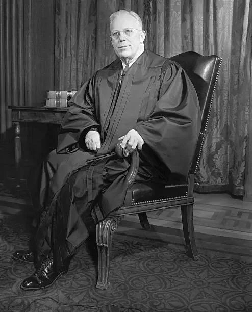 A seated man in judicial robes poses confidently in a formal setting, with a serious expression and a wooden chair accenting the dignified ambiance.
